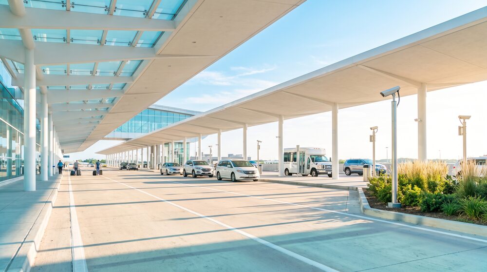 An airport terminal departures curb in daylight with cameras mounted along the median